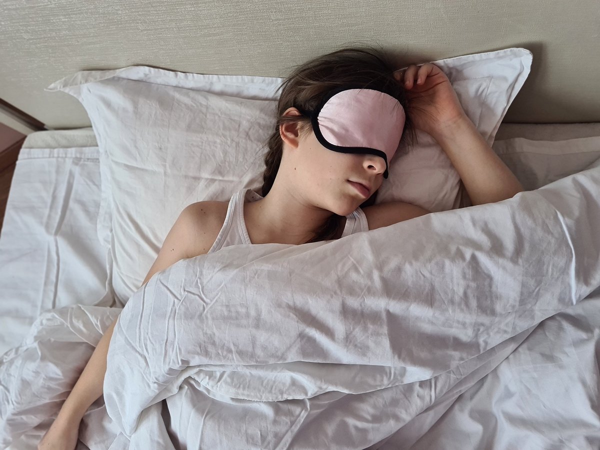 A young girl is sleeping peacefully in a bed with white sheets and a pink sleep mask covering her eyes