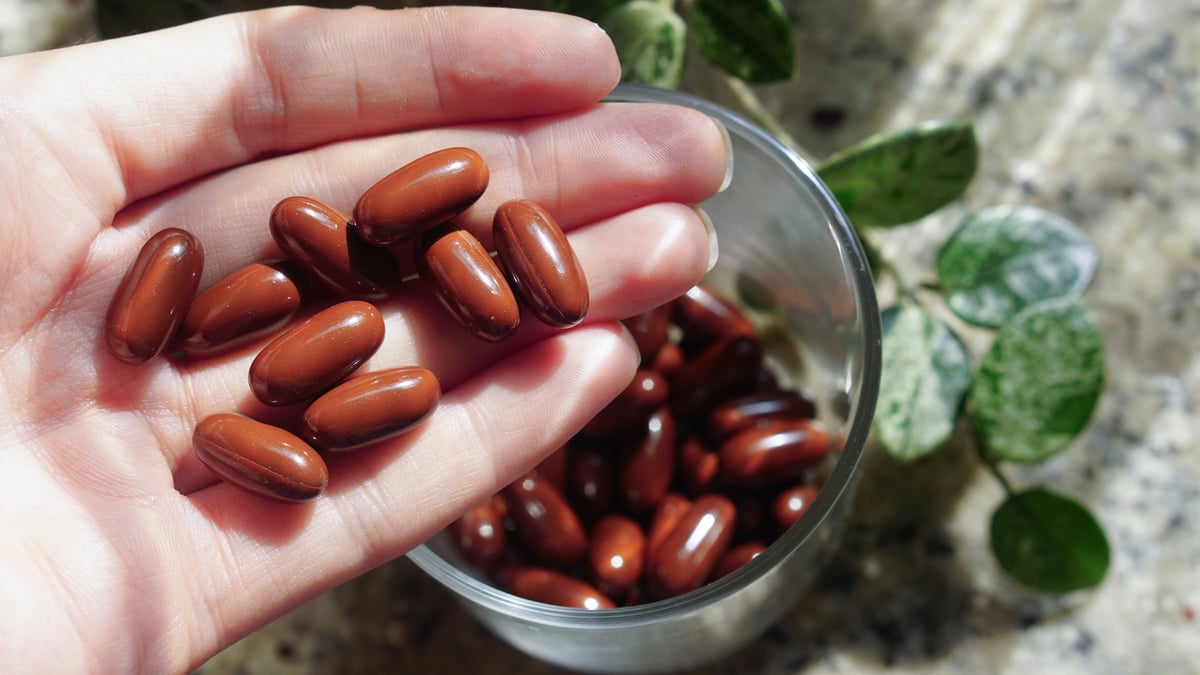 a hand holding several brown oval capsules. Below the hand is a clear glass jar filled with similar capsules, placed on a speckled countertop. A green leafy plant appears in the background, adding a natural touch.