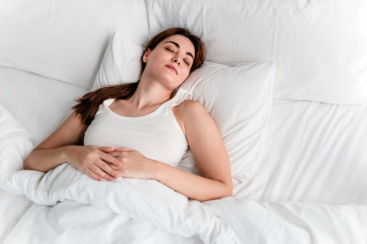 A young woman rests peacefully on a bed with white sheets in a bright, cozy bedroom, capturing a serene moment of sleep, relaxation, and wellness