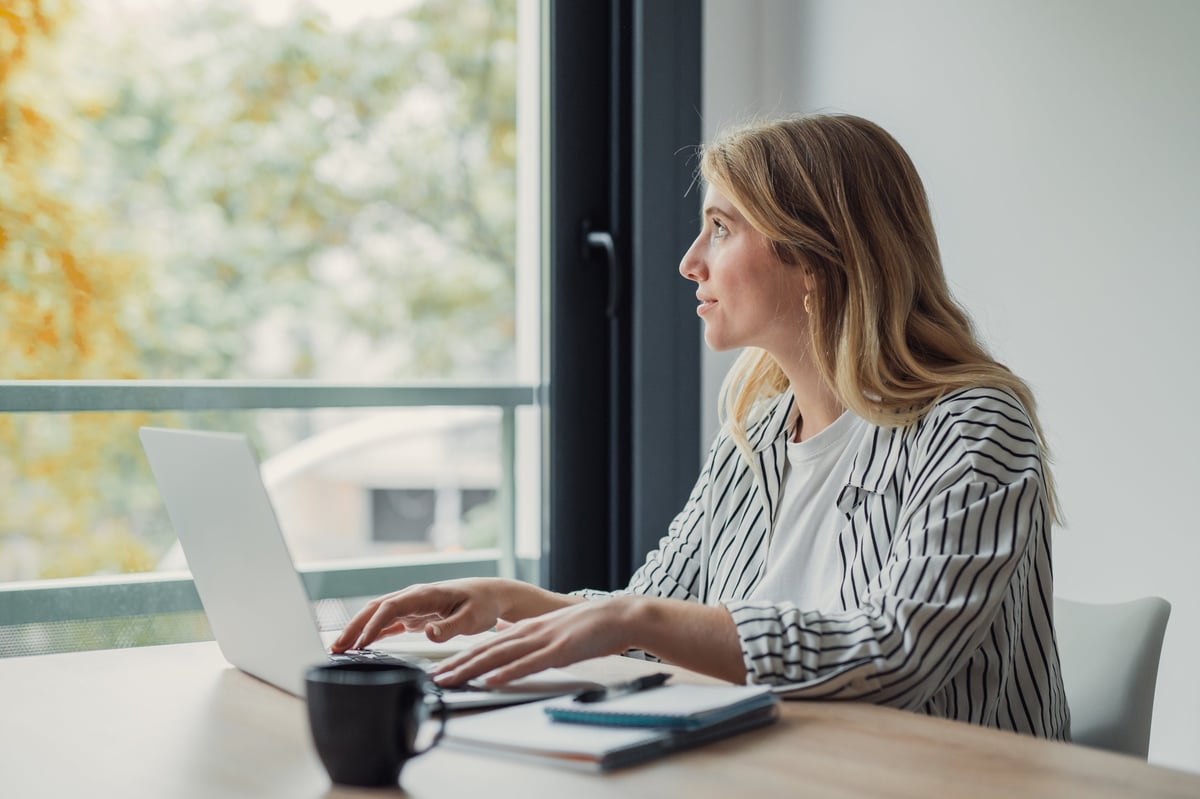 Young woman working remotely on a laptop at a desk by a window, pausing to gaze outside in thoughtful reflection during a daylight break in her home office routine
